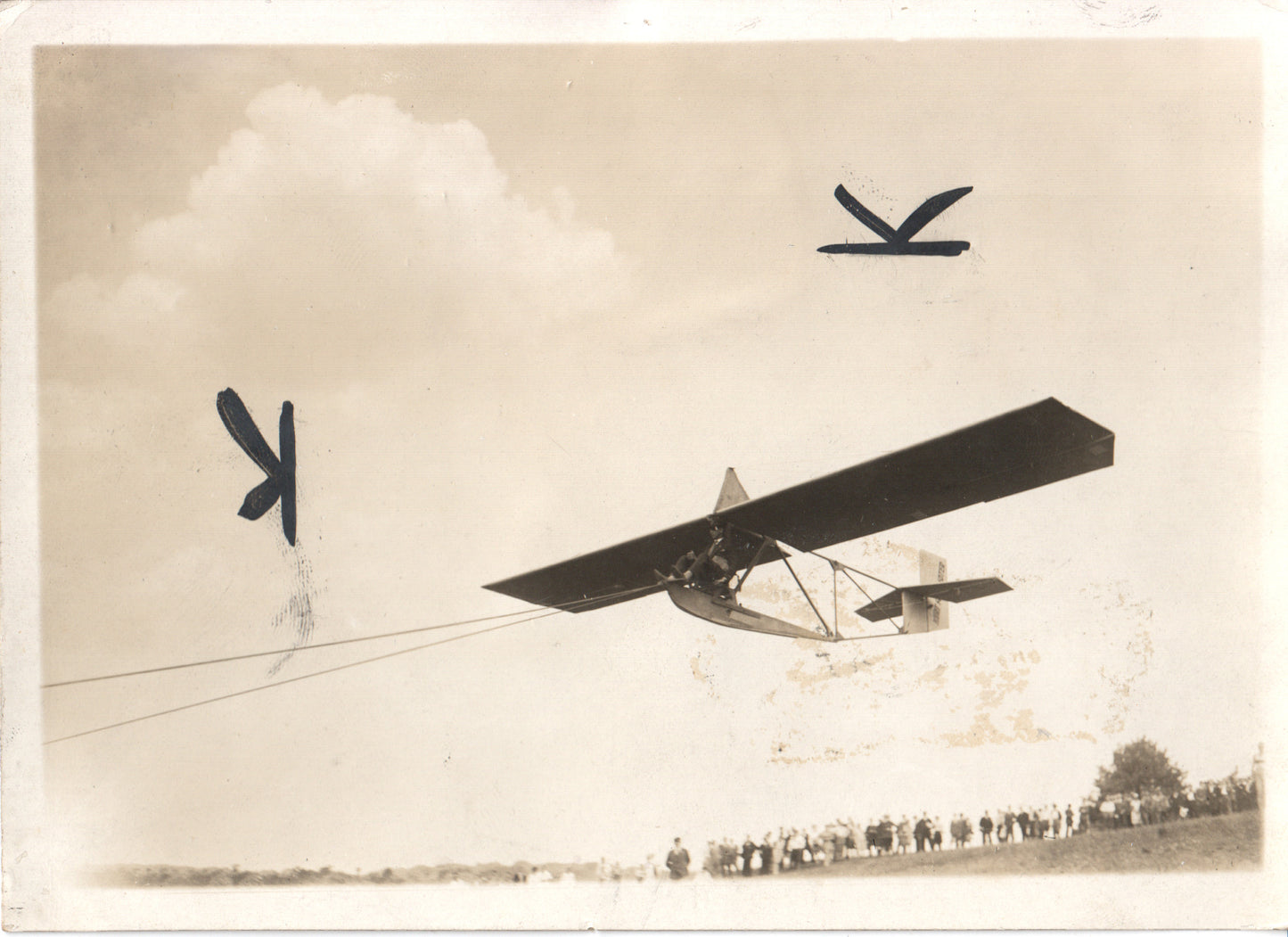 A vintage photograph of an aerial glider in flight, with a crowd watching below. - Image: Folder24-23
