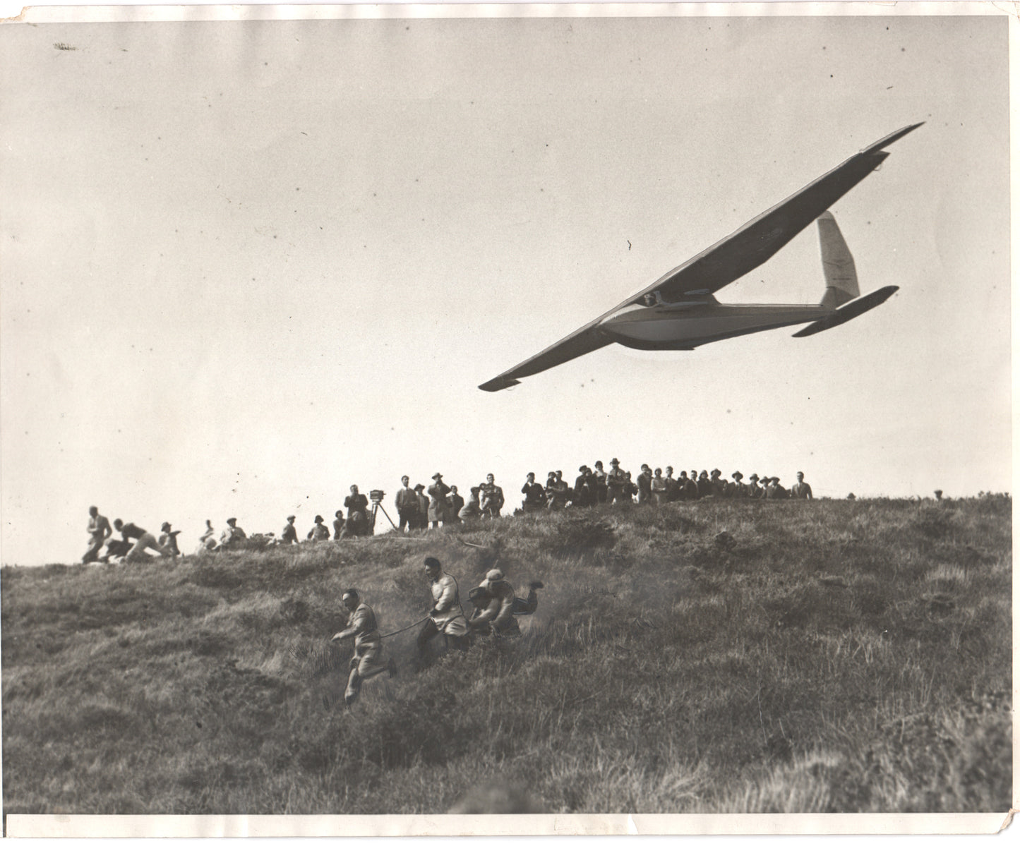 Vintage black and white photograph of a glider aircraft in flight with a crowd watching. - Image: Folder16-23