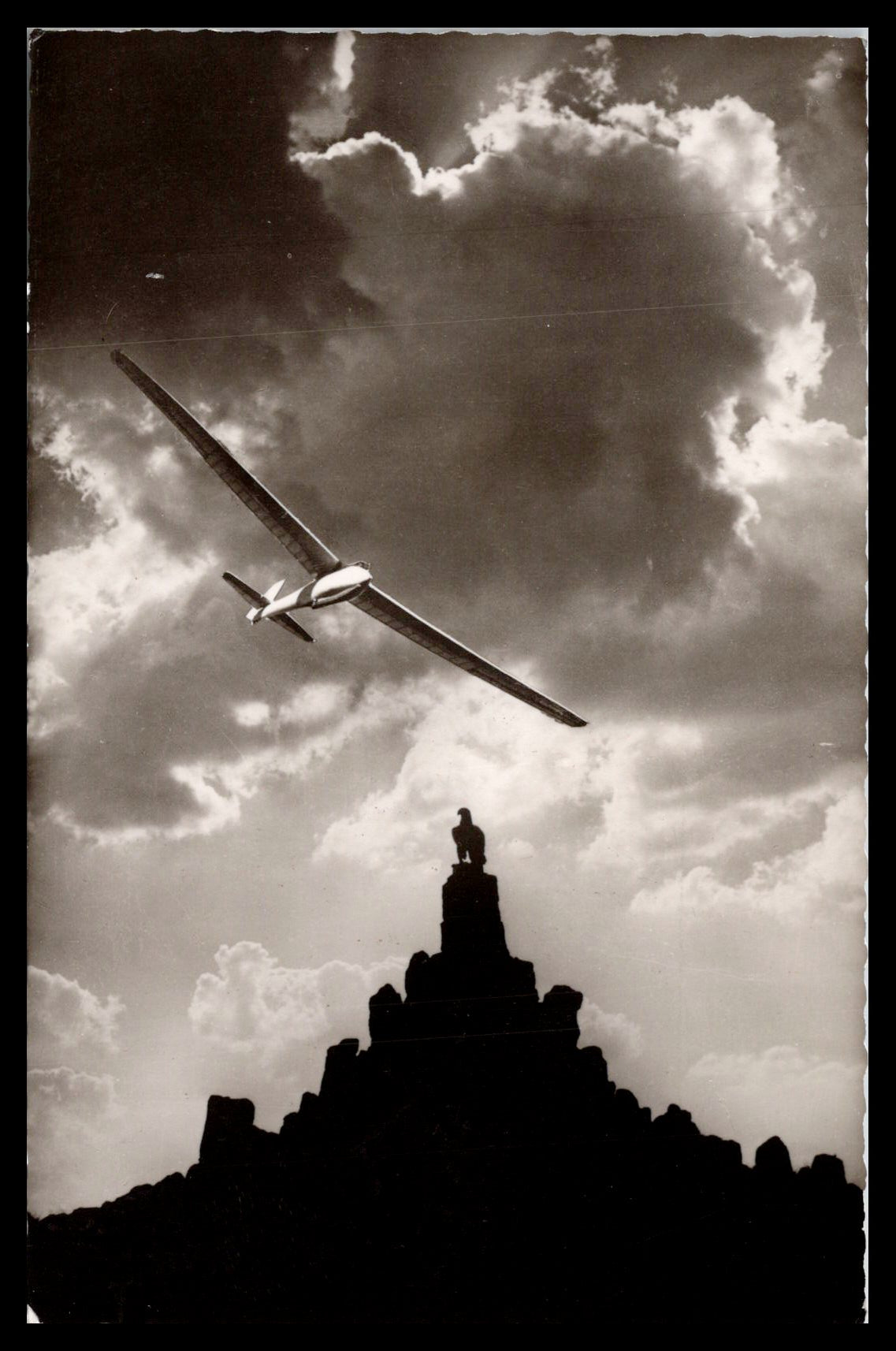 Black and white photo of a glider flying above an eagle statue on a rocky pedestal. - Image: MA-PC_0033