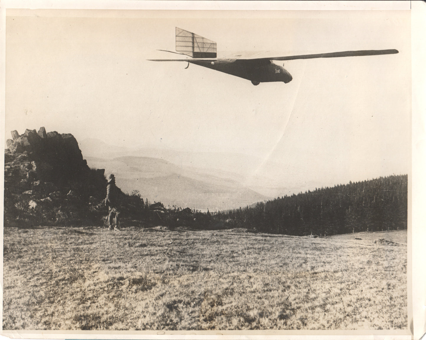 Black and white photograph of a glider flying over a scenic landscape with a figure and a dog in the foreground. - Image: Folder3-1