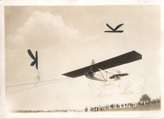 A vintage photograph of an aerial glider in flight, with a crowd watching below. - Image: Folder24-23