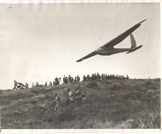 Vintage black and white photograph of a glider aircraft in flight with a crowd watching. - Image: Folder16-23