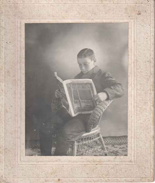 Black and white photograph of a young boy sitting in a chair, reading a magazine. - Image: 2025-04-13-0060