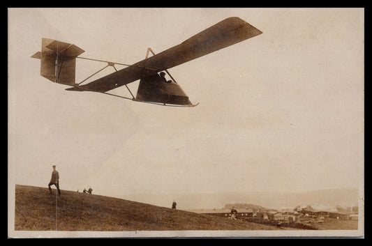 Vintage photograph of a model aircraft flying over a grassy hill with spectators in the background. - Image: MA-PC_0027