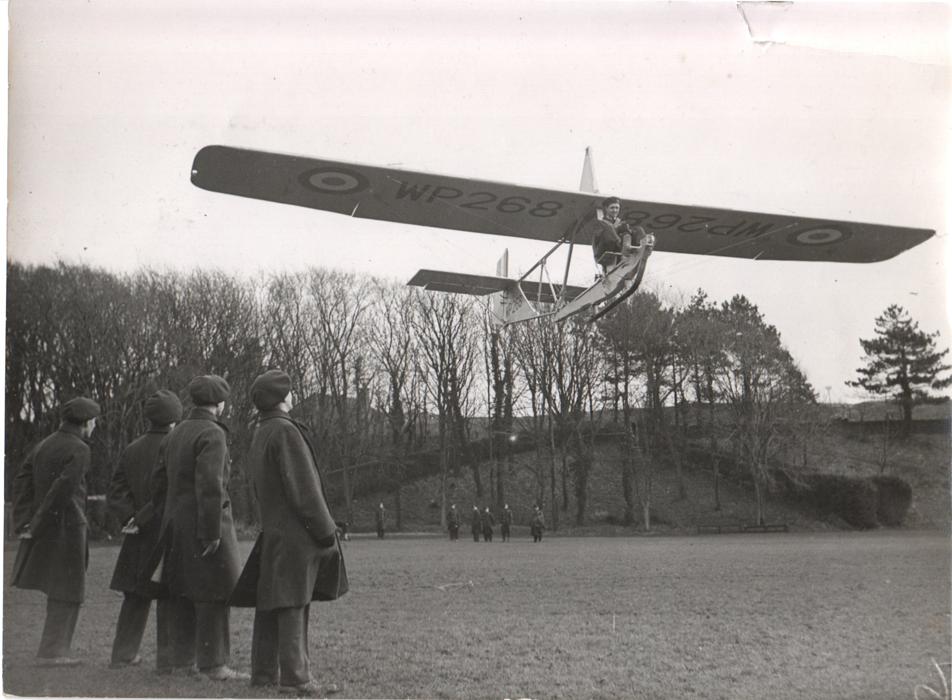 A vintage photograph of a WWII-era British training aircraft flying above a group of observers. - Image: Folder27-17