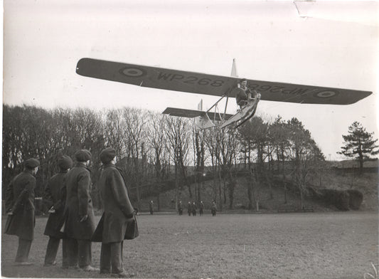A vintage photograph of a WWII-era British training aircraft flying above a group of observers. - Image: Folder27-17