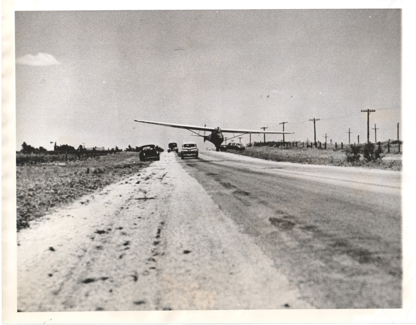 A vintage black and white photograph of a plane landing on a rural road with parked cars. - Image: Folder17-7