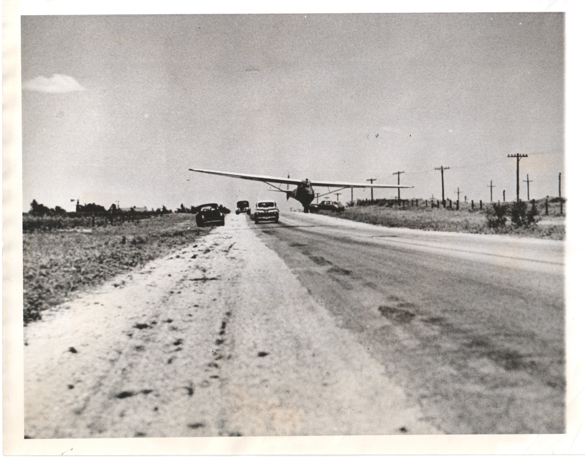 A vintage black and white photograph of a plane landing on a rural road with parked cars. - Image: Folder17-7