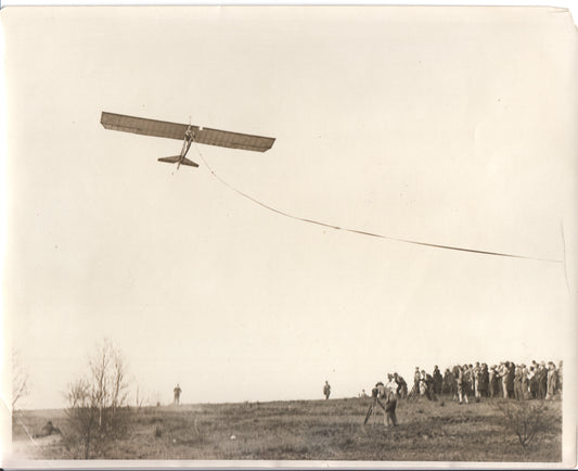 Black-and-white photograph of a vintage aircraft in flight over a crowd on a hillside. - Image: Folder22-8