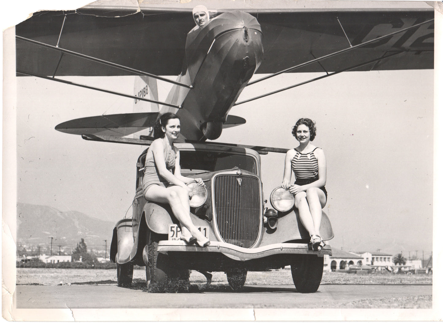 Vintage photograph of two women in bathing suits sitting on a classic car with an airplane above them. - Image: Folder28-1
