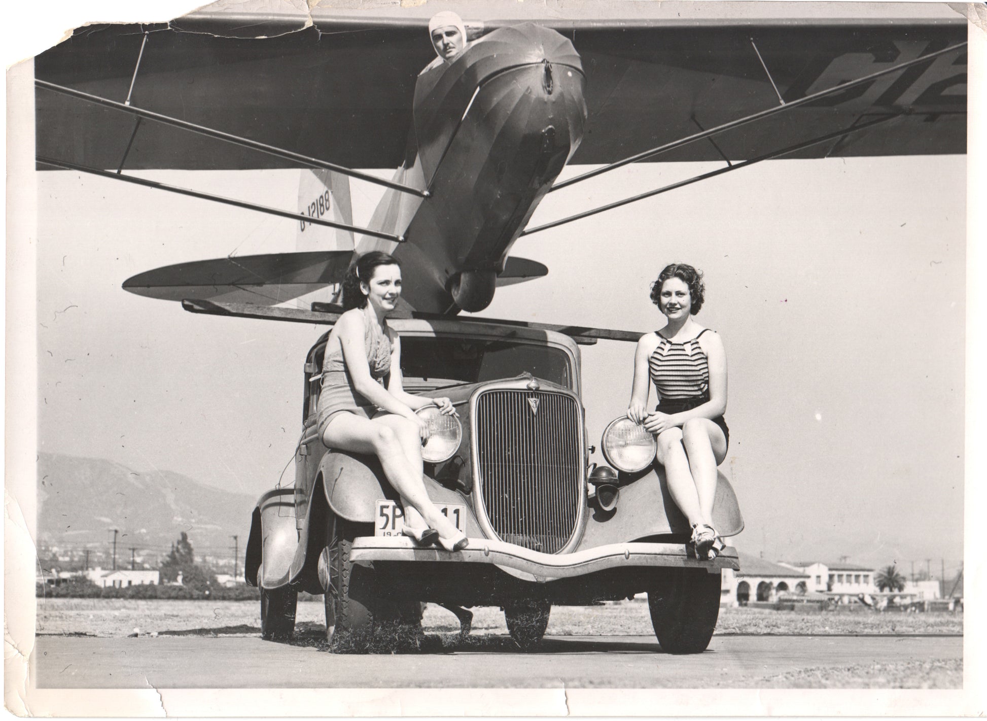Vintage photograph of two women in bathing suits sitting on a classic car with an airplane above them. - Image: Folder28-1