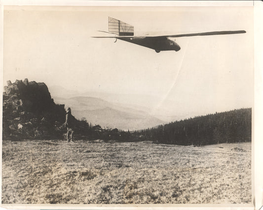 Black and white photograph of a glider flying over a scenic landscape with a figure and a dog in the foreground. - Image: Folder3-1