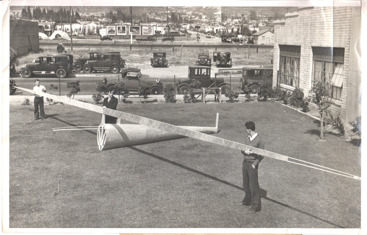 Vintage black-and-white photograph of young men with a glider model and classic cars in the background. - Image: Folder3-5